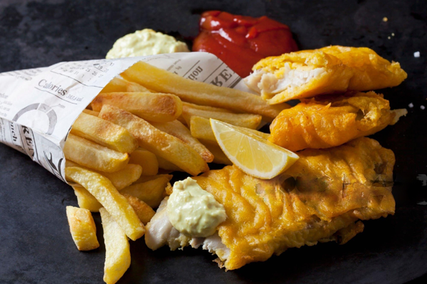 A classic fish and chips meal on a dark surface, featuring battered fish fillets, a lemon wedge, tartar sauce, ketchup, and thick-cut fries served in a newspaper-print paper cone.