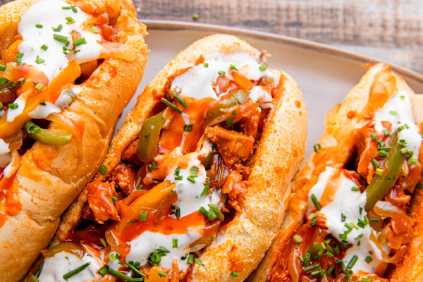 A trio of sub sandwiches or fajita rolls filled with chopped meat, sautéed peppers, and onions, topped with a drizzle of white creamy sauce and fresh chives.